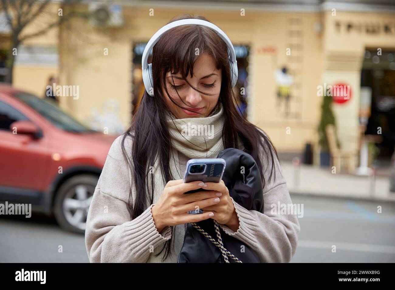 Happy concentrated young woman in earphones using smartphone outdoor. Freelance, influencer, chatting concept. Stock Photo