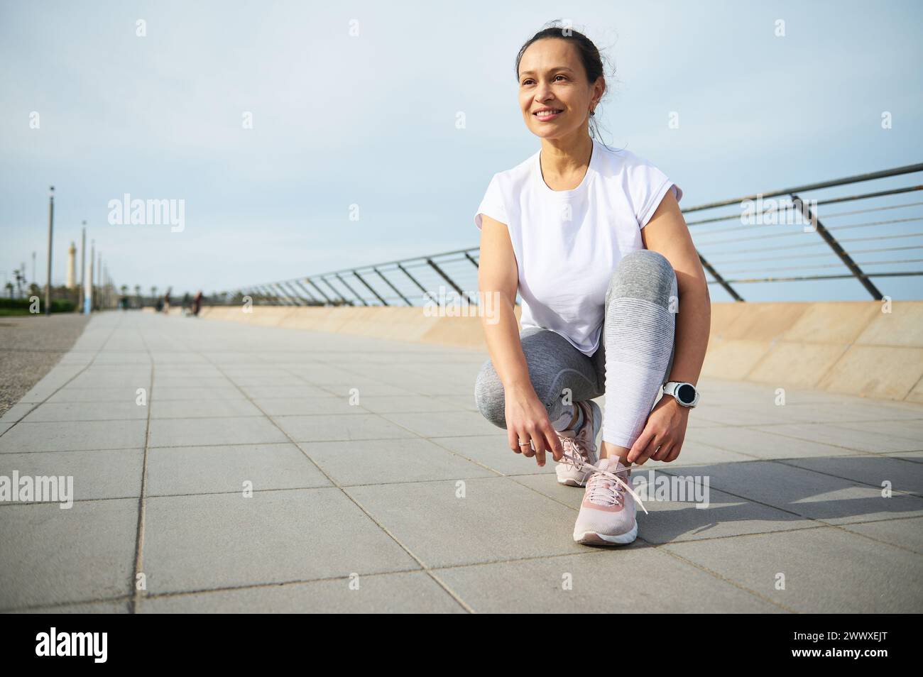Confident young woman runner tying laces on running shoes, ready for jog on Atlantic promenade on beautiful sunny summer day. Pretty female athlete lo Stock Photo