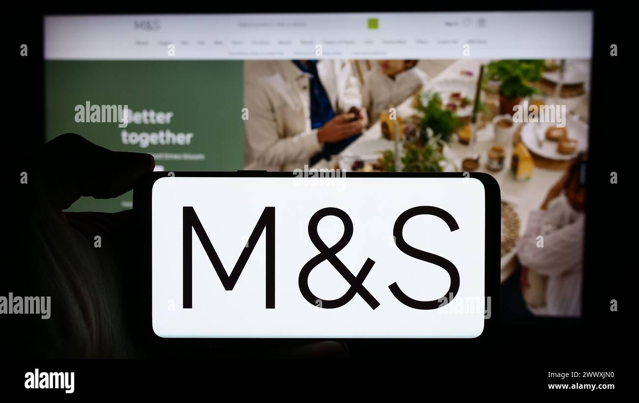 Person holding cellphone with logo of British retail company Marks and Spencer Group plc in front of business webpage. Focus on phone display. Stock Photo