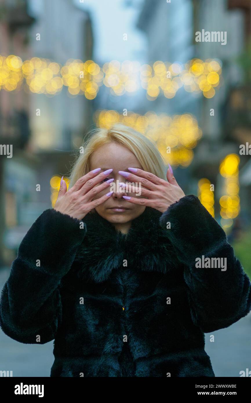 A blonde woman in a black coat covers her eyes, with evening bokeh lights behind her Stock Photo