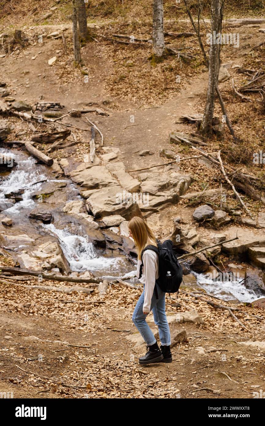 Young blond trekker with travel backpack, walking near forest stream enjoying views Stock Photo