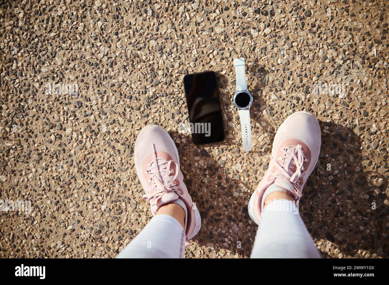Top view feet of a sportswoman in pink running sports shoes, a smart wrist watch and smartphone with black blank mockup digital screen on the asphalt. Stock Photo
