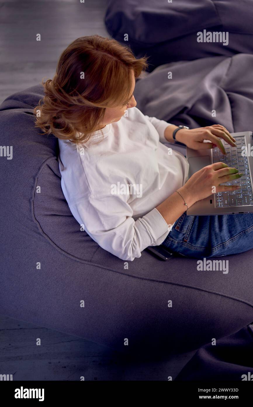 woman in casual clothes is sitting at a laptop on bean bag in front of ...