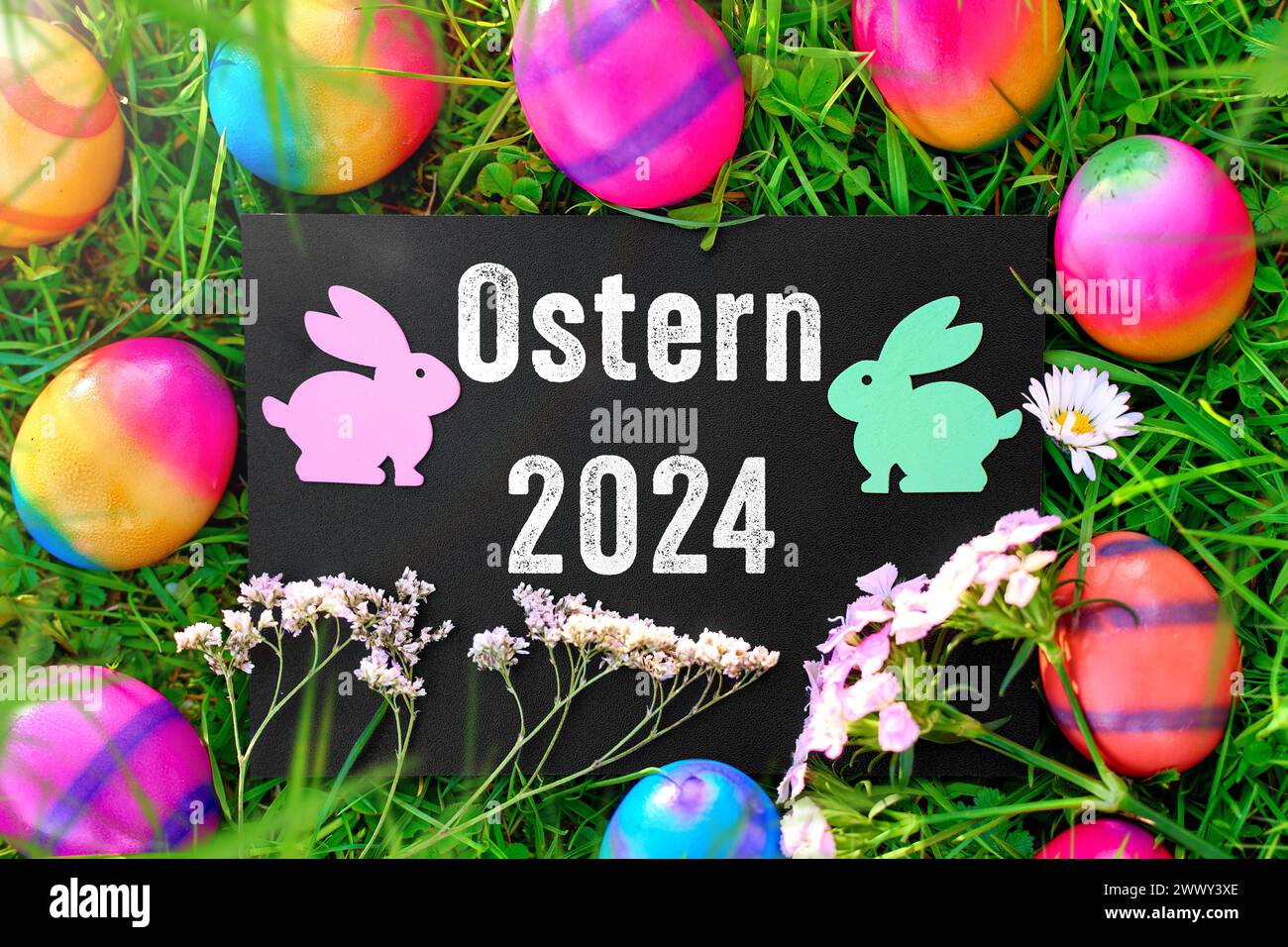 26 March 2024: Easter 2024, lettering on a board in a green meadow with colorful Easter eggs and Easter bunny figures. PHOTOMONTAGE *** Ostern 2024, Schriftzug auf einer Tafel in einer grünen Wiese mit bunten Ostereiern und Osterhasen Figuren. FOTOMONTAGE Stock Photo