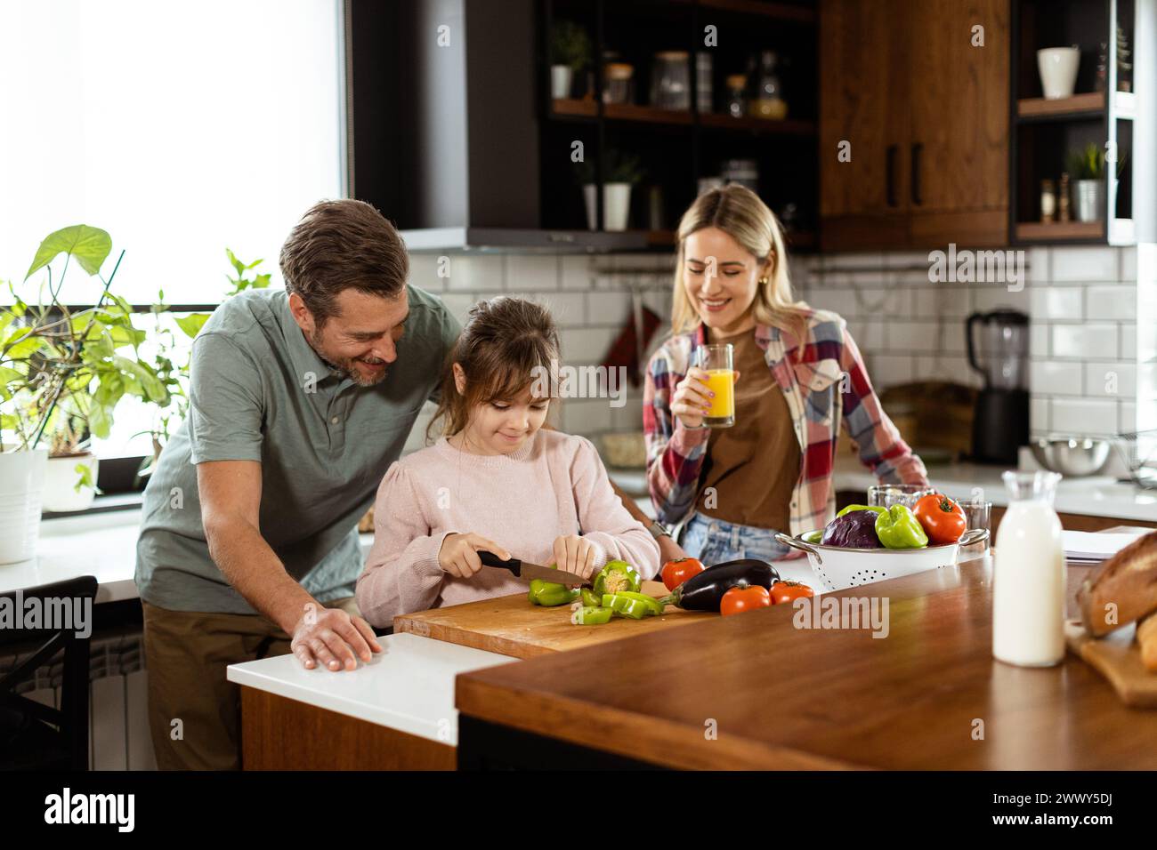 A family prepares a meal together, with dad helping daughter cut vegetables and mom enjoying a juice. Stock Photo
