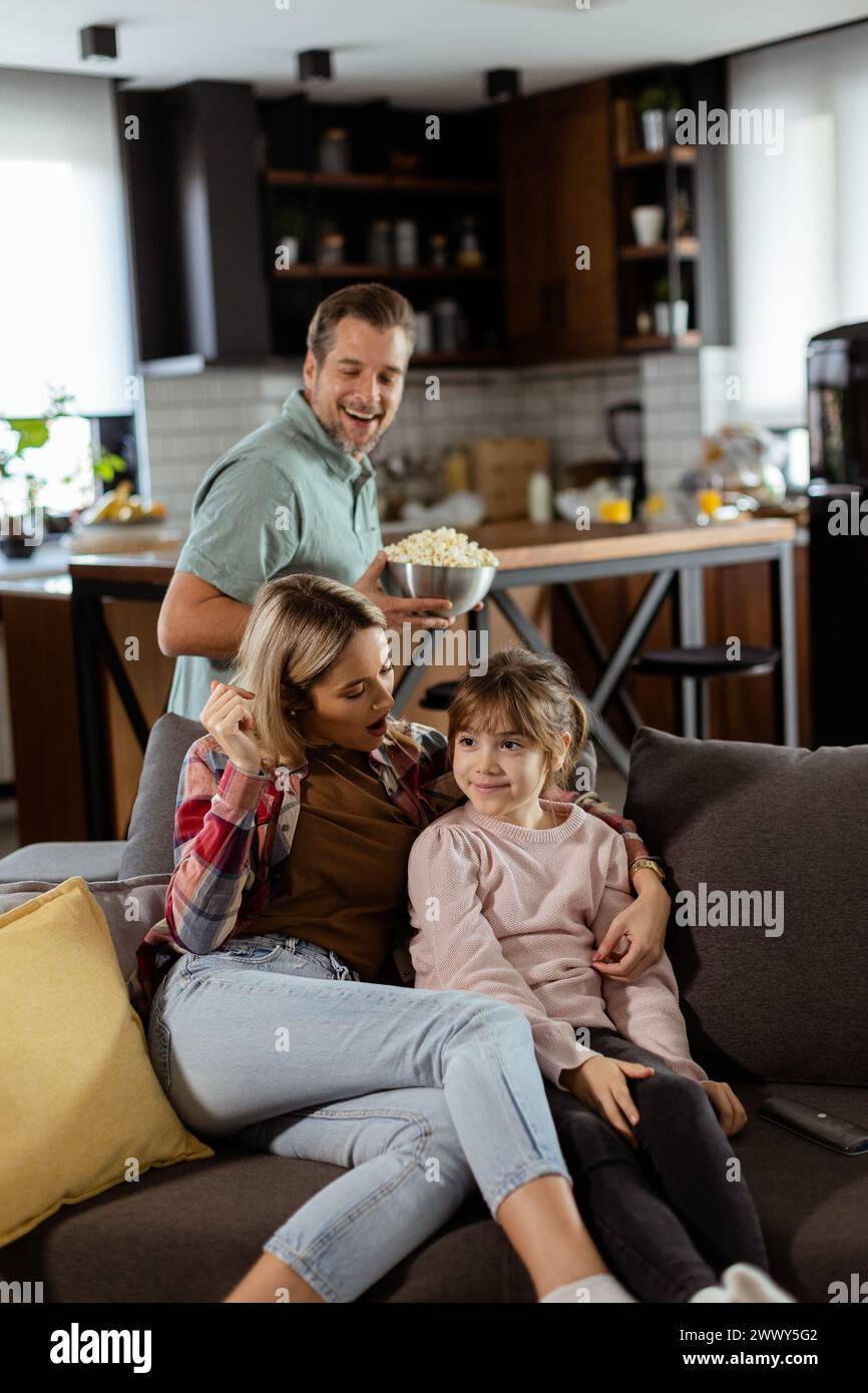 A family of three is comfortably nestled on a couch, their faces reflecting excitement and attentiveness as they share a bowl of popcorn during a susp Stock Photo