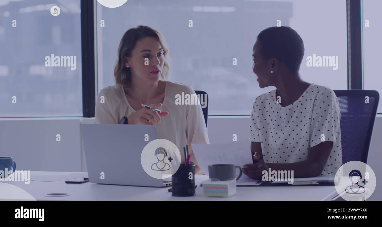 Image of people icons floating over diverse female colleagues working on laptop in office. global business, digital interface and technology concept d Stock Photo