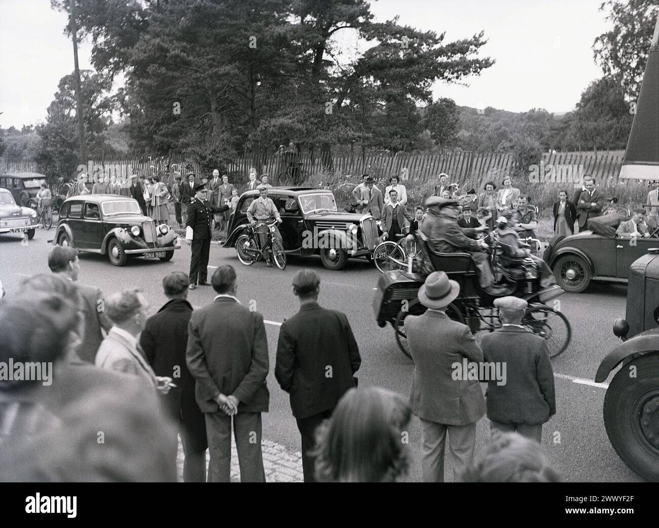 1950s, historical, spectators roadside watching a veteran car drive off on a vintage car run, with a British policeman standing in the middle of the road signaling the other road users to wait, England, UK. Stock Photo