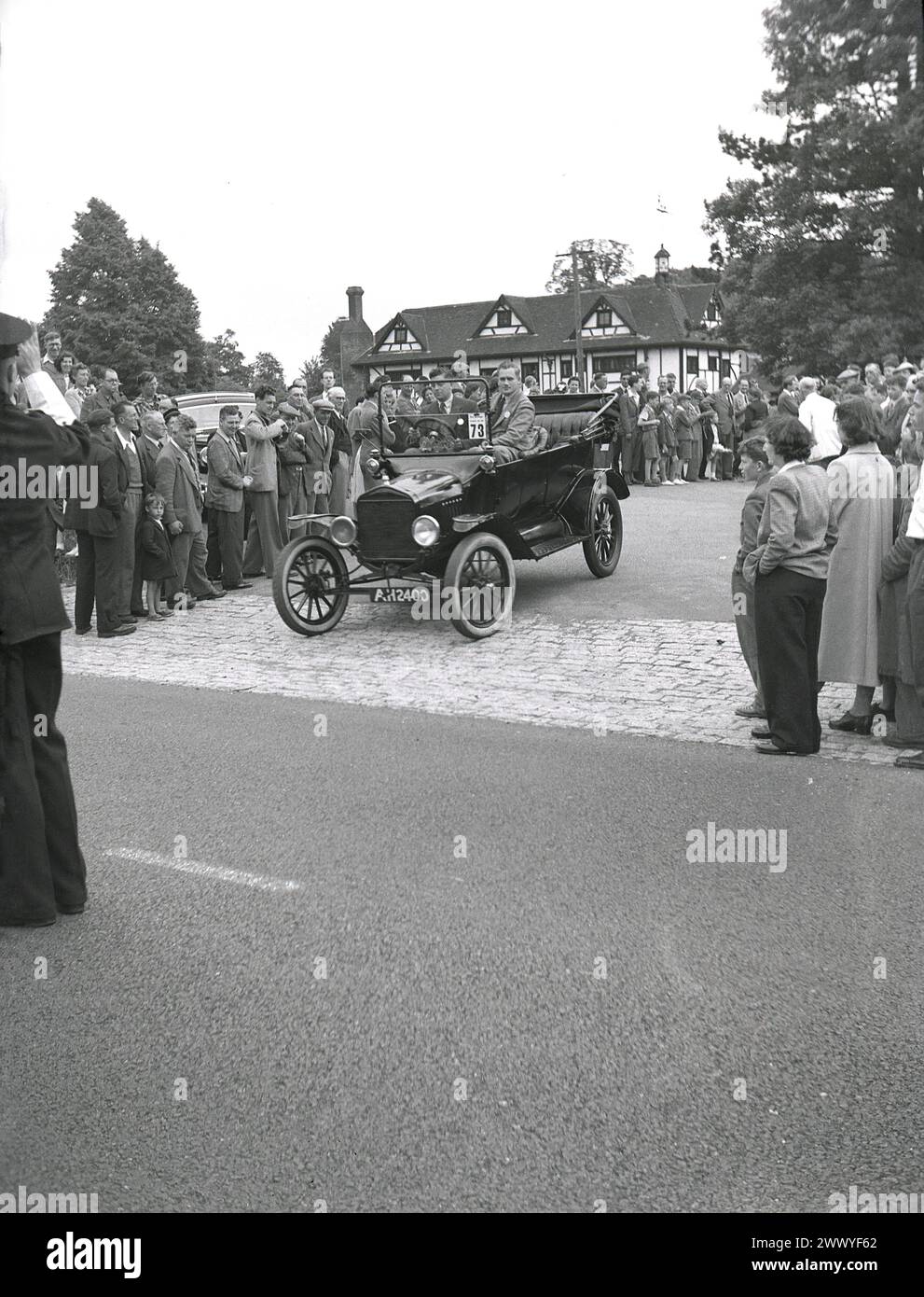 1950s, historical, a crowd watching a veteran car - open-top, registration AH 2400 - drive off on a vintage car run, with a British policeman standing in the middle of the road signaling for the other road users to wait, England, UK. Motor cars built before WW1 are generally considered veteran cars, although the strict interpretation is that only cars built pre 1904 can be called veteran cars and that cars built between 1905-1918 should be referred to as Edwardian cars. Cars made beween 1919 and 1930 are generally classified as Vintage. Stock Photo