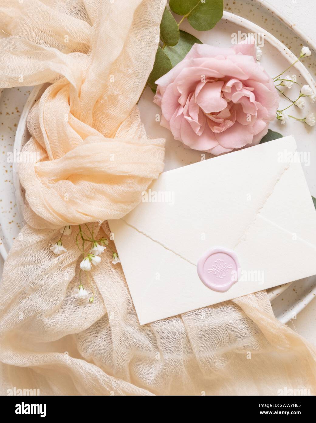 Sealed envelope near cream tulle fabric knot and light pink roses on plates top view, copy space. Wedding stationery mockup. Romantic table place with Stock Photo