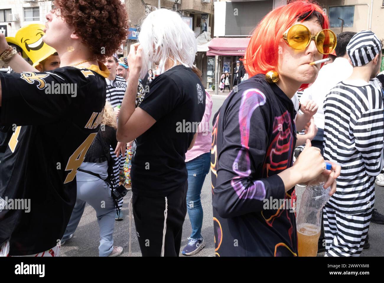 Bnei Brak, Israel. 24th Mar, 2024. A young man wearing an orange wig and sunglasses, smokes a cigarette during the Purim celebrations. Ultra-Orthodox Jews Celebrate Purim in Bnei Brak, Israel. The holiday commemorates the salvation of the Jews in ancient Persia from a plot to annihilate them. A joyous holiday, it is celebrated by both secular and nonsecular Jews, most notably by dressing up in costumes and drinking, according to the Talmud, 'until they cannot distinguish between ''˜cursed is Haman' and ''˜blessed is Mordechai. (Credit Image: © Syndi Pilar/SOPA Images via ZUMA Press Wire) E Stock Photo