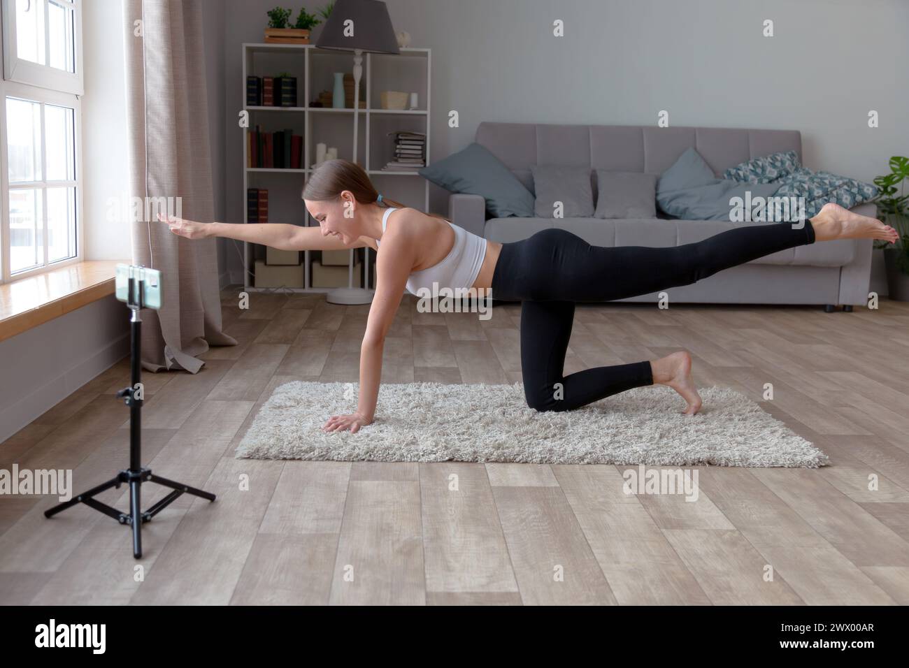 woman in black leggings and a white top does yoga at home on the carpet, recording an online fitness video for her audience with a smartphone standing Stock Photo
