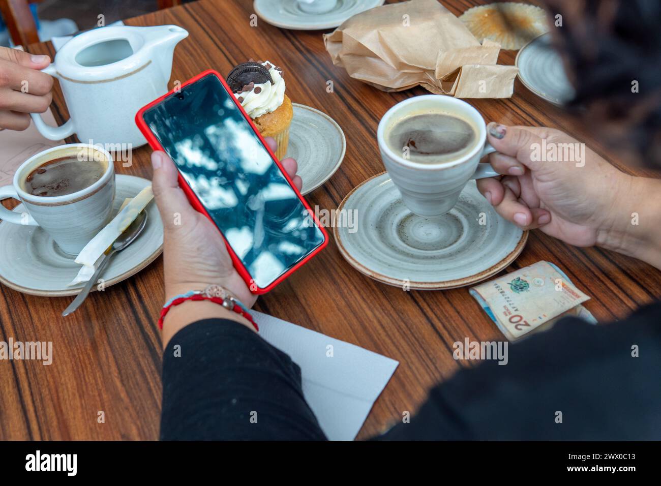 Unrecognizable woman drinking a cup of coffee while looking at her cell phone next to a cupcake and a 20 Colombian peso bill in a cafeteria Stock Photo