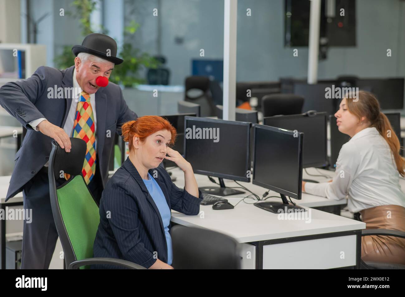 An elderly Caucasian man in a clown costume amuses two Caucasian women in the office.  Stock Photo