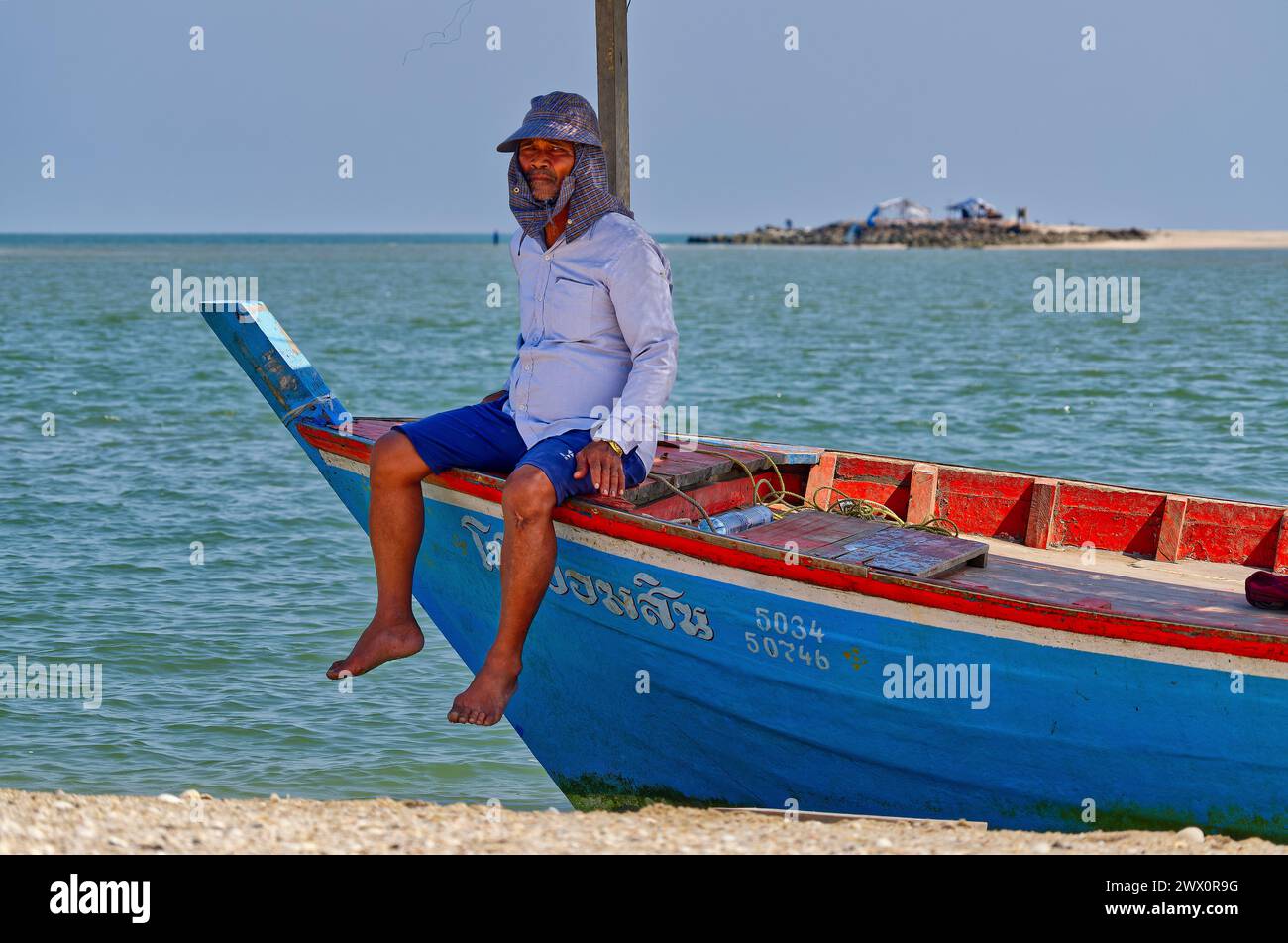 Fisherman resting on blue and red boat landed on a sand spit in the Gulf of Thailand Stock Photo