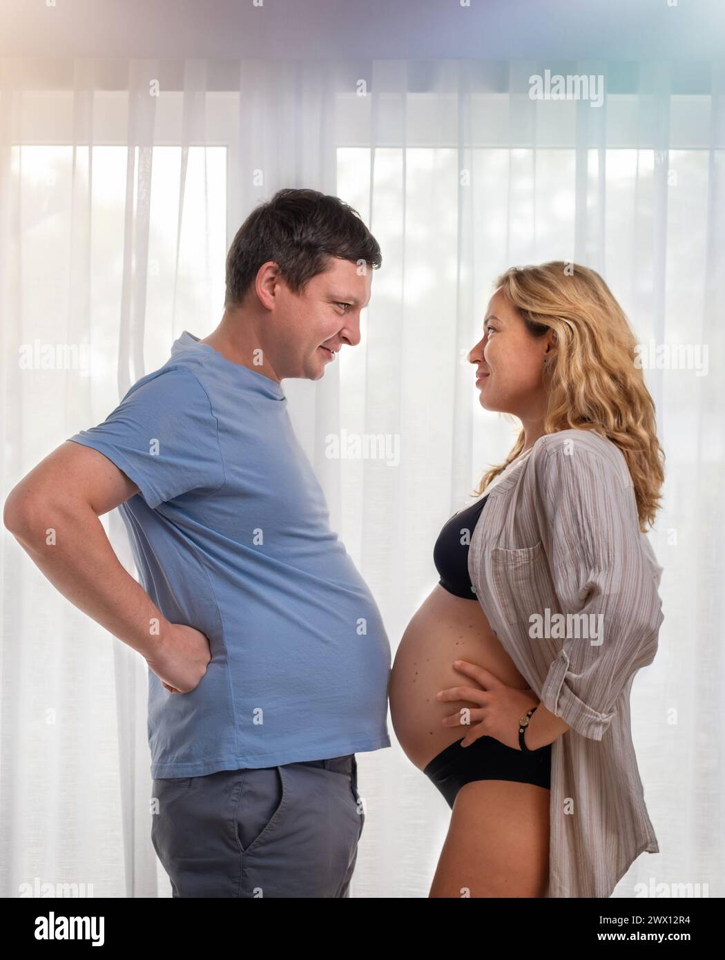A pregnant girl and her husband measure their bellies near the window Stock Photo