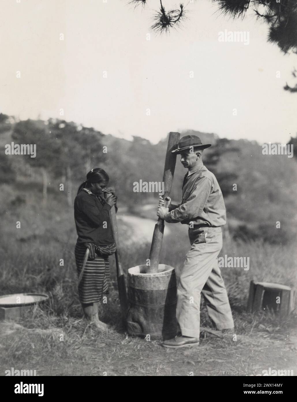 Taken at Camp John Hay Native Barrio, Baguio, Philippine Islands An Ifugao woman watches an American Soldier try his hand at pounding rice ca. 1931 Stock Photo