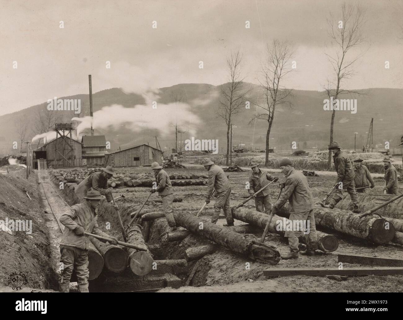 Soldiers loading logs on car showing sawmill in the distance. Men of