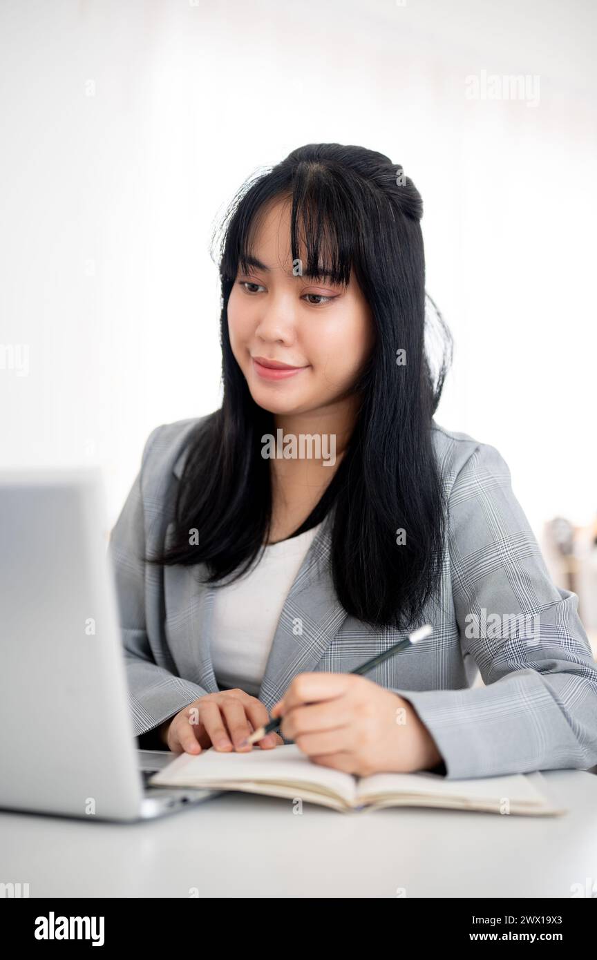 A young Asian businesswoman or female office worker is looking at her laptop screen while taking notes in the notebook, working in a modern office. Stock Photo