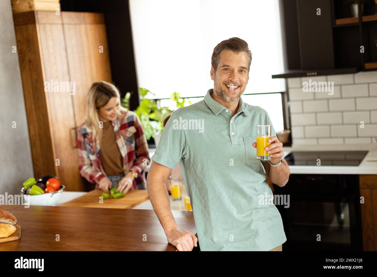 Smiling man holds a glass of juice while two women cook in a bright kitchen Stock Photo
