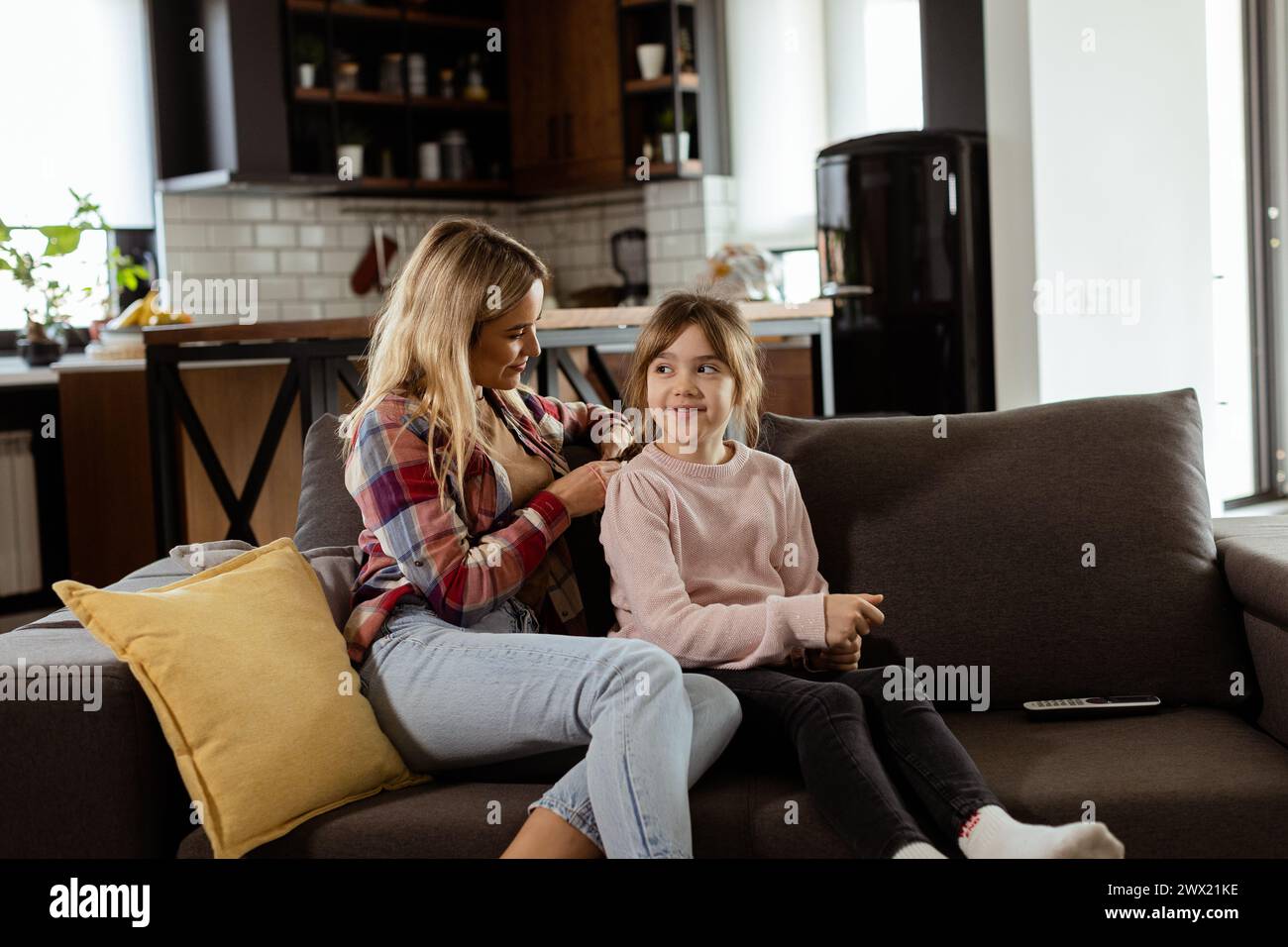 Gentle touch enriches a quiet morning as a mother intricately weaves her daughters hair on the living room sofa Stock Photo