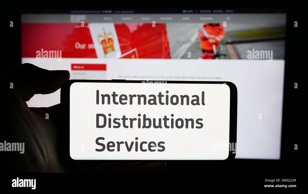 Person holding smartphone with logo of British company International Distributions Services plc in front of website. Focus on phone display. Stock Photo