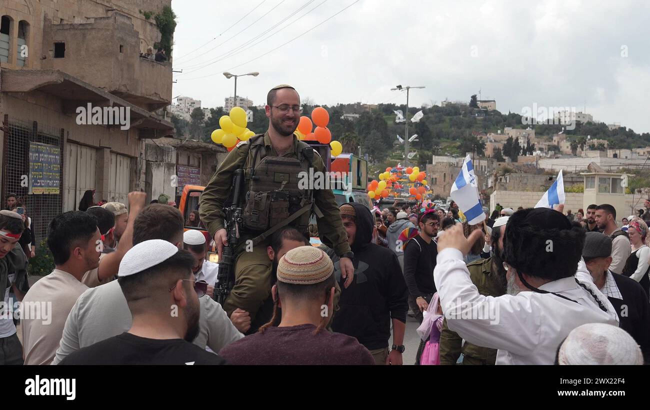 Jewish settlers wearing costumes dance with an Israeli soldier as they march down Al-Shuhada street, which is largely closed to Palestinians, during the annual Purim parade on March 24, 2024 in Hebron, Israel. Stock Photo