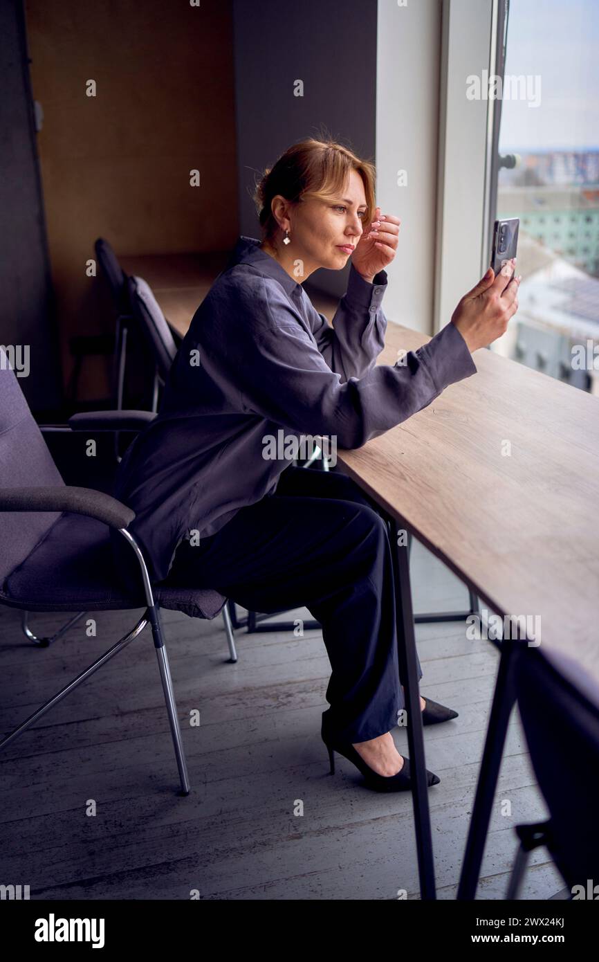 beautiful middle age business woman wearing gray shirt, wide leg pants and black stilettos fix her hair looking into phone camera in modern workspace Stock Photo
