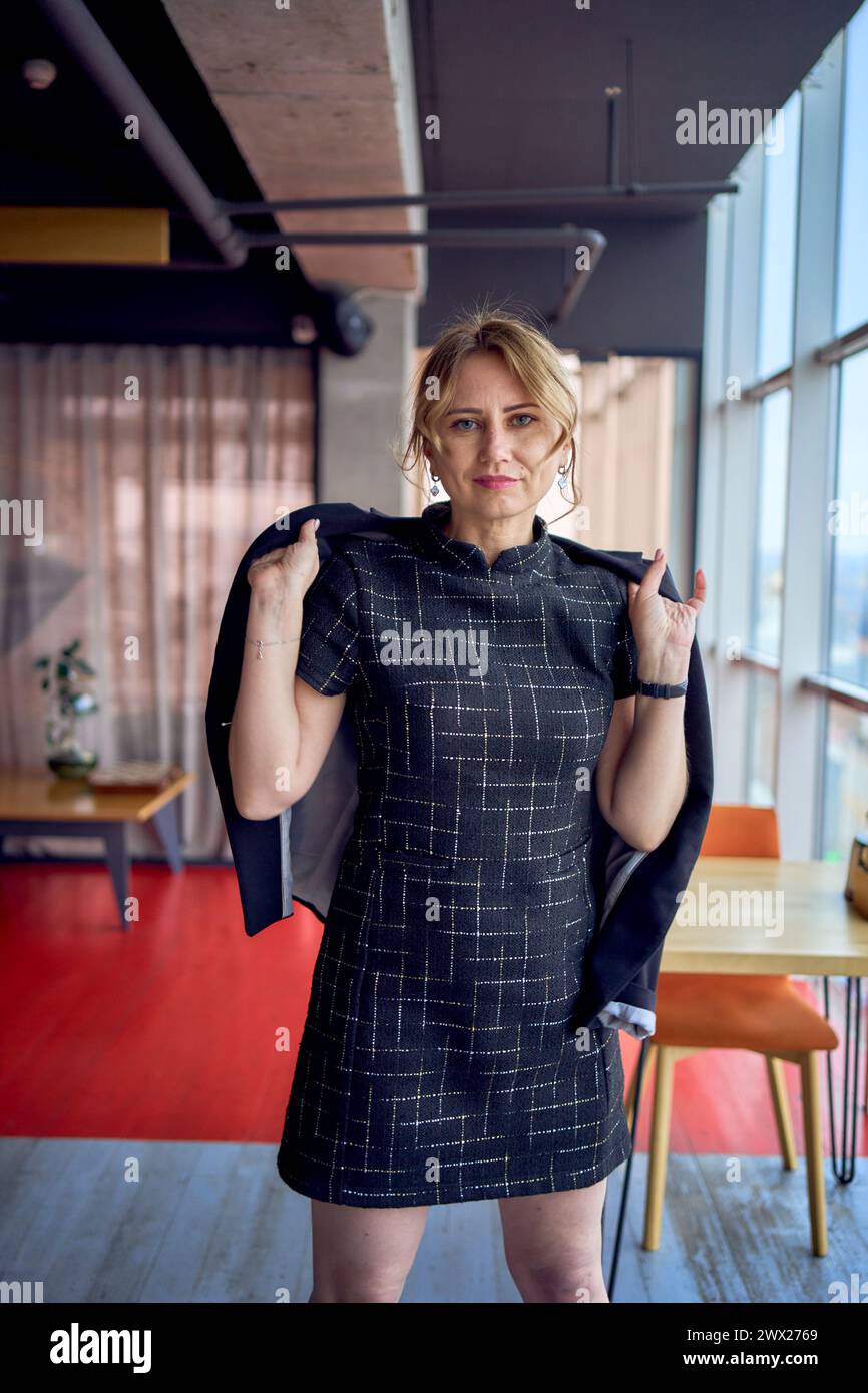 an  attractive woman in a black business style mini dress throws a jacket over her shoulders in a modern coworking space with panoramic windows and a Stock Photo