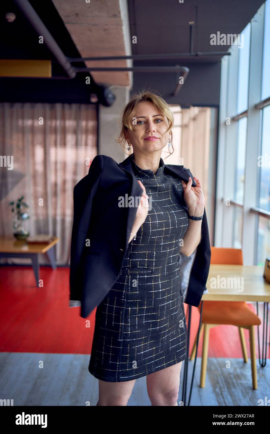 an  attractive woman in a black business style mini dress throws a jacket over her shoulders in a modern coworking space with panoramic windows and a Stock Photo