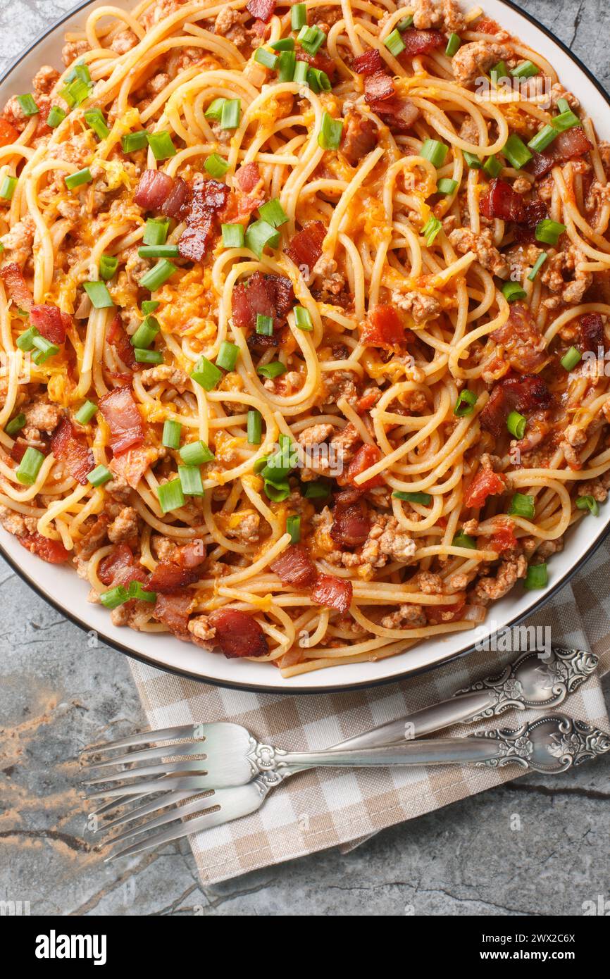 American style Spaghetti pasta with bacon, minced meat, cheddar cheese, onion and spicy tomato sauce close-up in a plate on the table. Vertical top vi Stock Photo