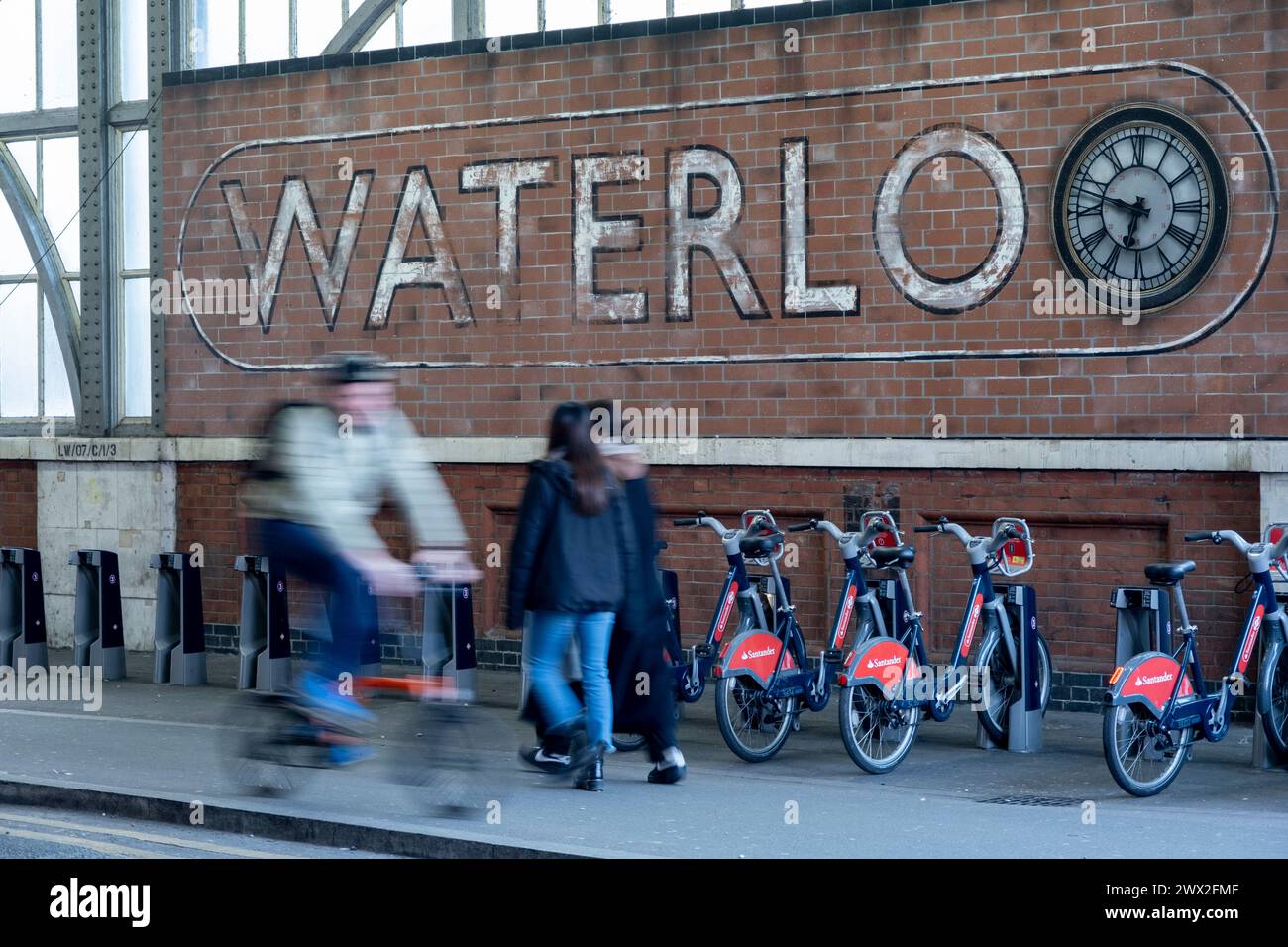 Faded sign for Waterloo Railway Station as people pass on bicycles on 6th March 2024 in London, United Kingdom. Waterloo station also known as London Waterloo, is a major central London terminus on the National Rail network in the London Borough of Lambeth. It is connected to a London Underground station of the same name. Waterloo is the third busiest station in the UK, handling close to 60 million passengers per year as of 2023. Stock Photo