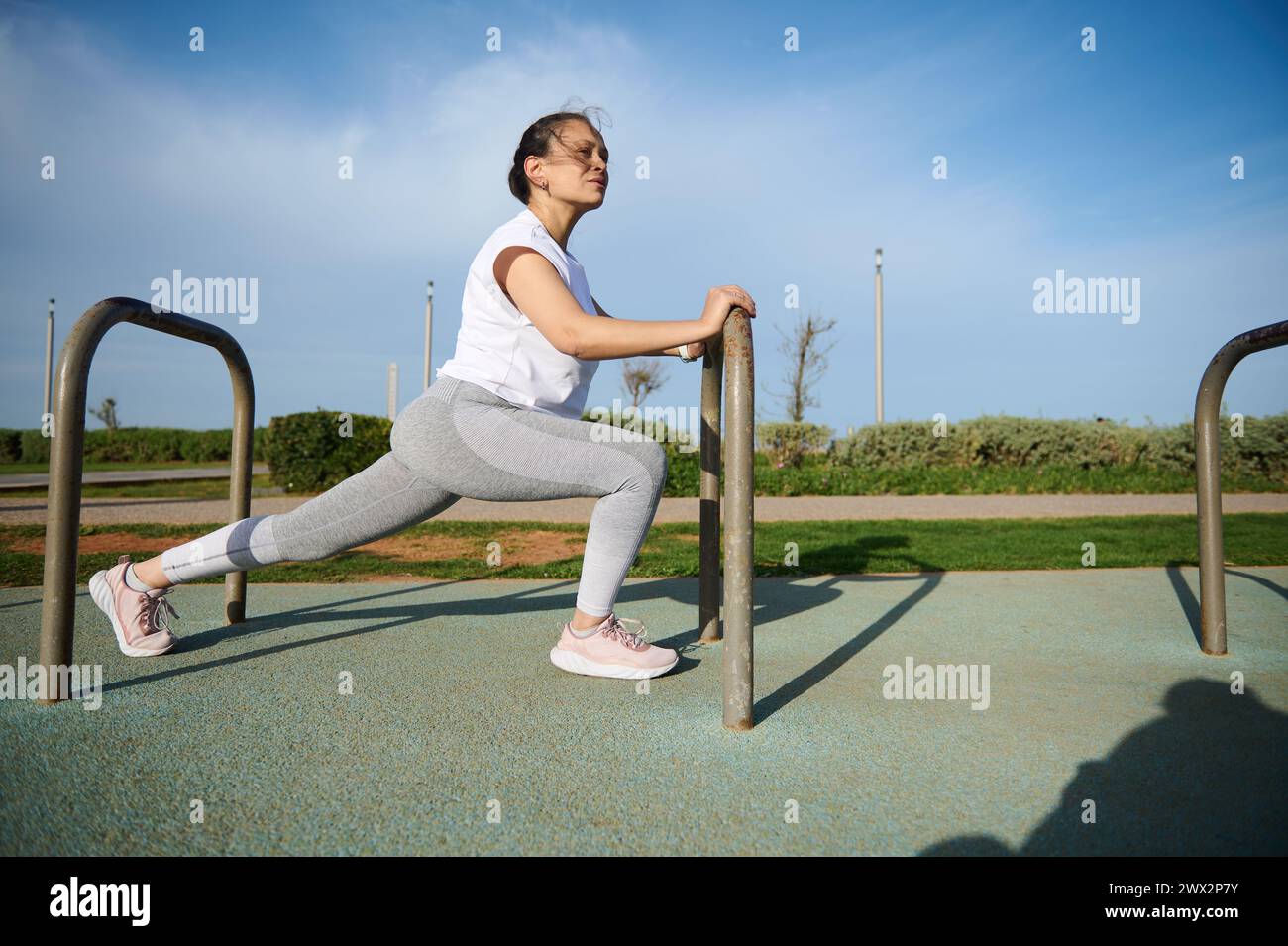 Full length portrait female athlete exercising with crossbar on the urban sportsground. Determined sports woman in white t-shirt and gray leggings, wa Stock Photo