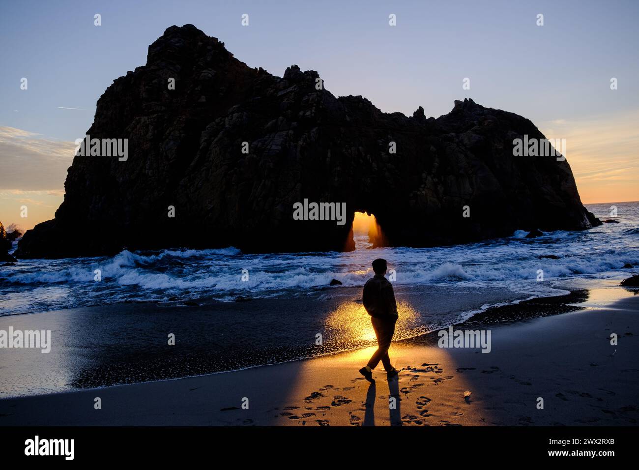 Sunset illuminates person in front of Window in the Rock feature of Keyhole Arch at Pfeiffer Beach in Big Sur on the Pacific Ocean in California, USA. Stock Photo