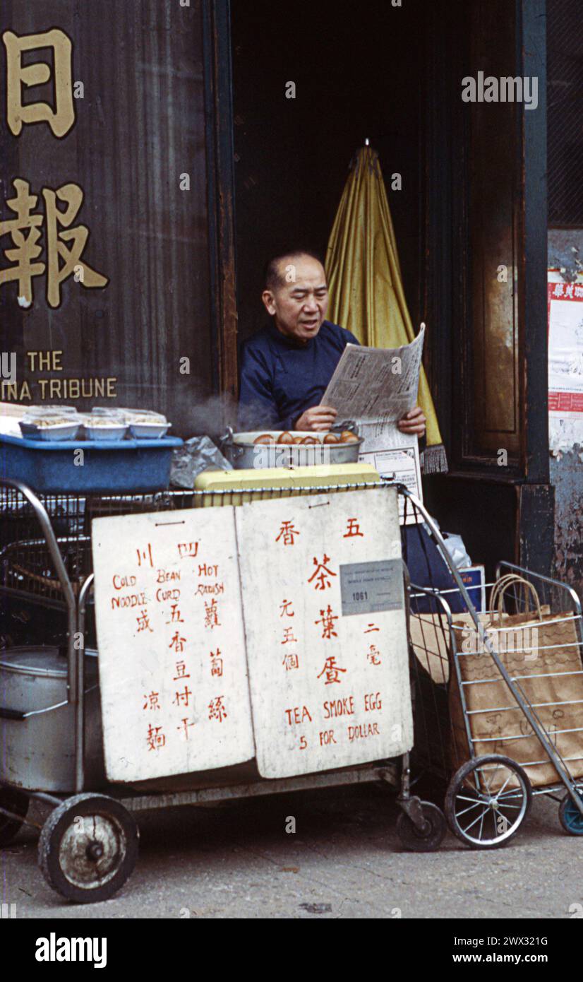 A Chinese American man reads a newspaper behind his stand where he's selling tea smoked eggs, cold noodles bean curd and hot radishes. On Canal Street in New York Chinatown in January 1979. Stock Photo