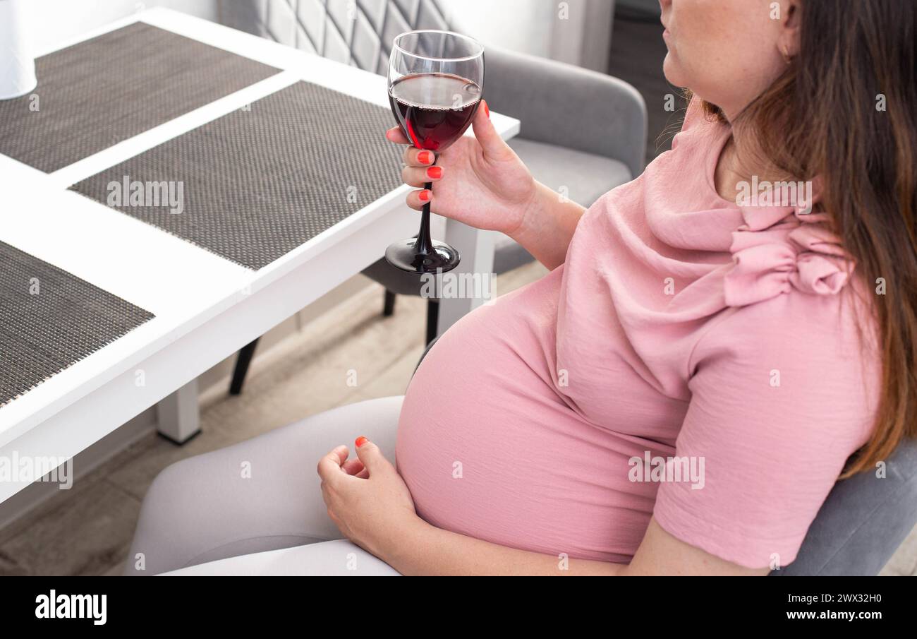 A pregnant girl with a big belly in a pink blouse holds a glass of wine. The concept of drinking alcohol during pregnancy, a bad habit. Stock Photo