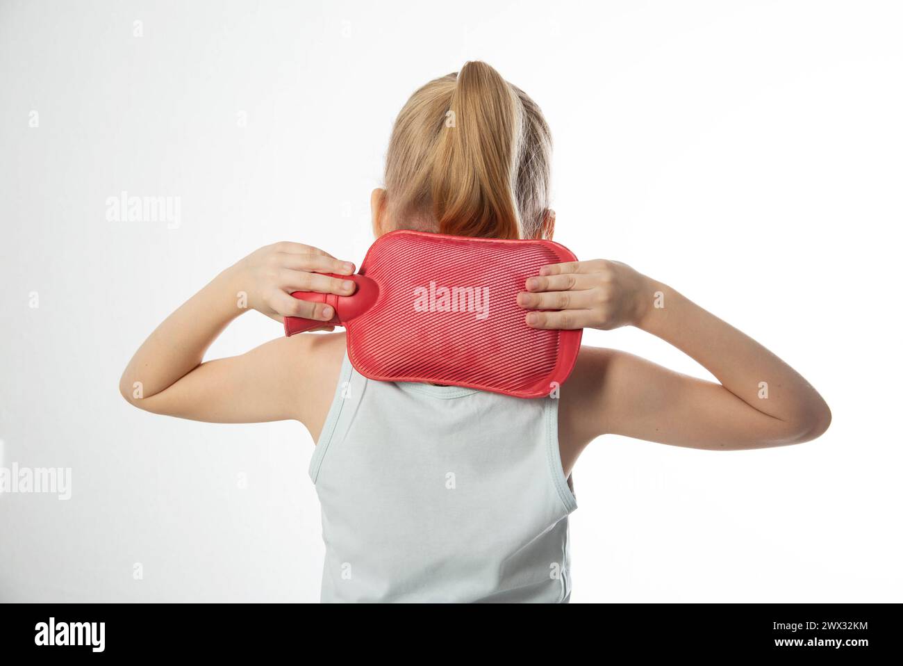 A little girl applies a red heating pad with warm water to her neck. Relieving neck pain with heat, muscle spasms. Stock Photo