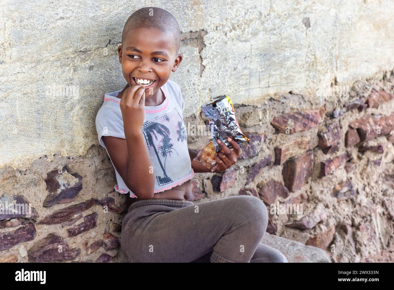 hungry single african girl with shaved head eating some snacks in front of the house in a village, Stock Photo