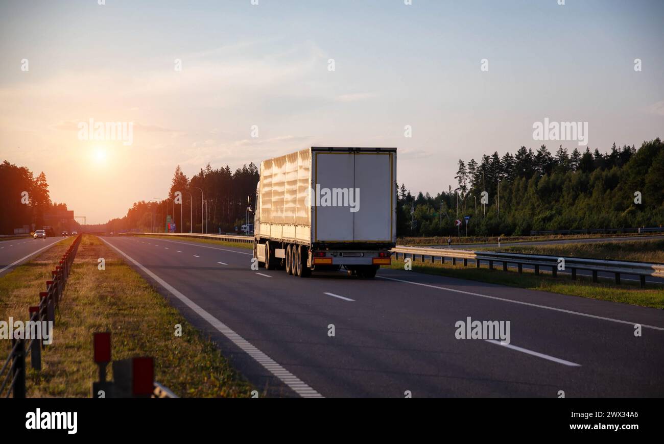 A truck with a tilt semi-trailer carries cargo along the highway in the evening against the backdrop of a summer sunset. The concept of cargo transpor Stock Photo
