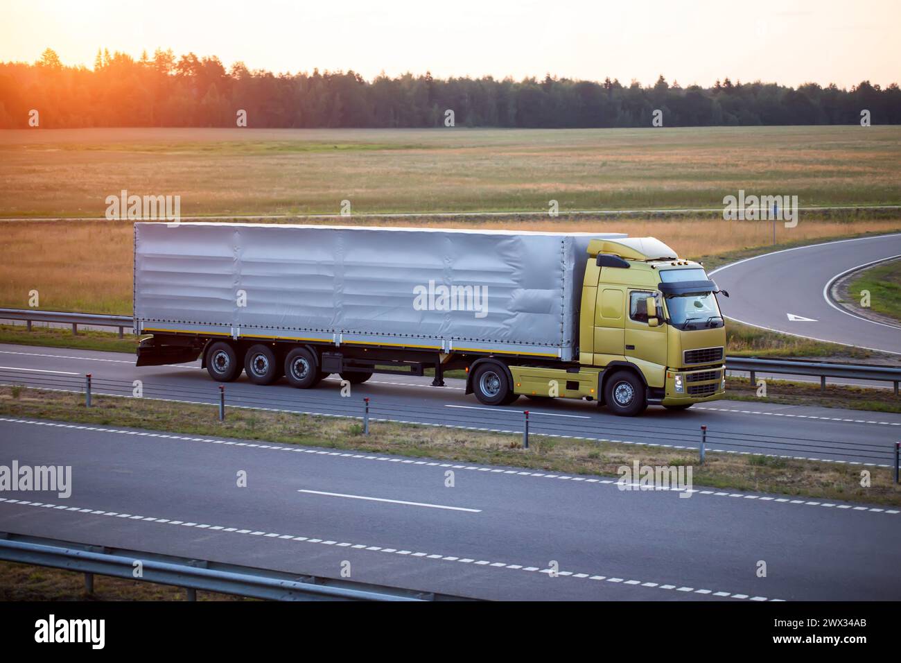 A truck with a tilt semi-trailer carries cargo along the highway in the evening against the backdrop of a summer sunset. The concept of cargo transpor Stock Photo