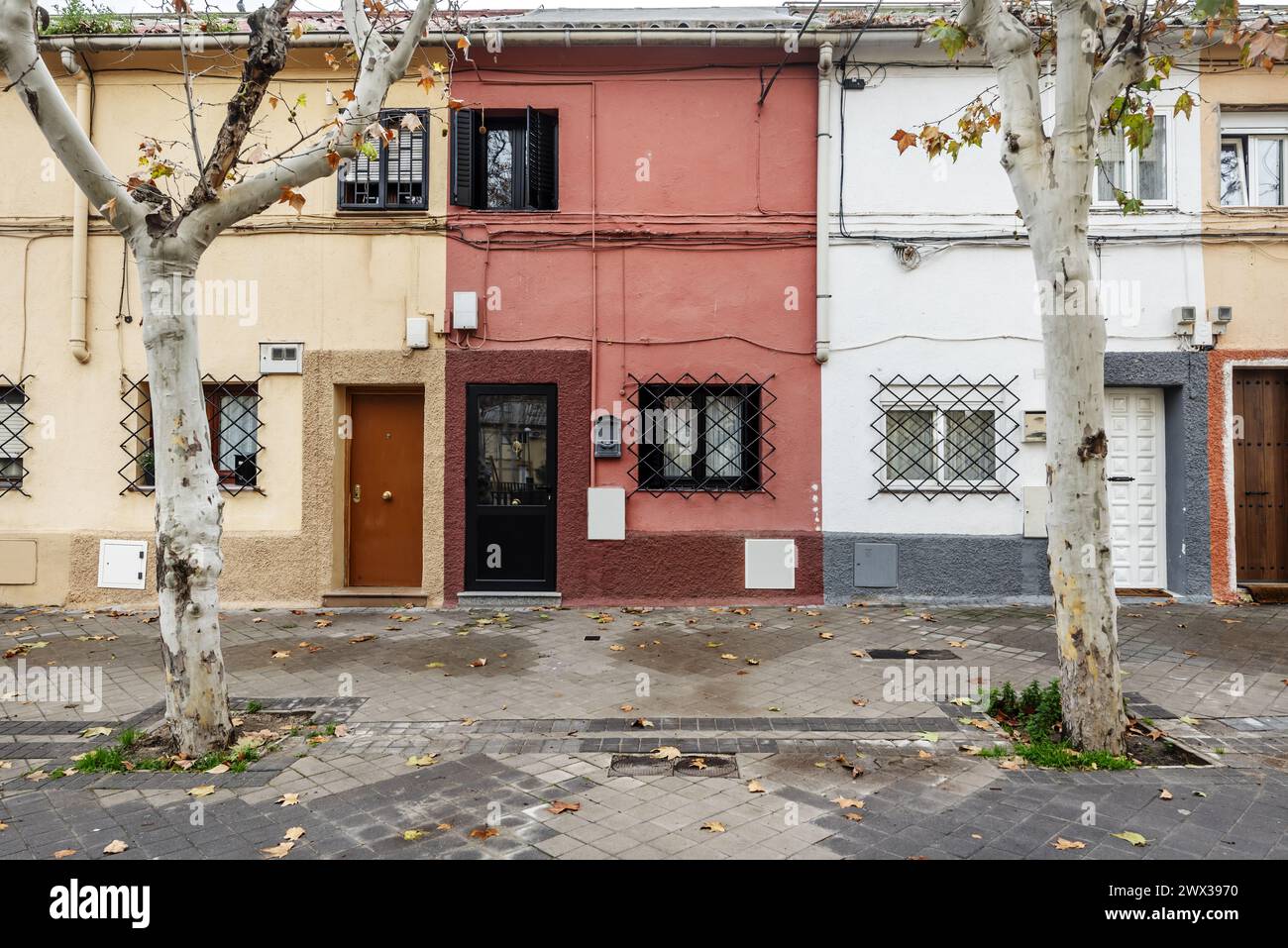 Facades of old houses of various colors of two heights with metal bars on the windows and trees with tree pits on the street Stock Photo