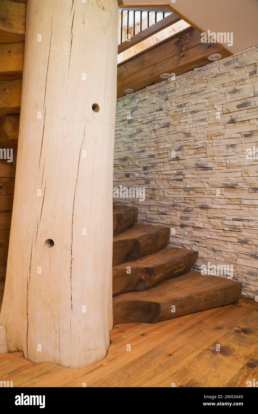 Twisting wooden staircase with brown stained half log steps attached to large bleached supporting central tree trunk post and tan natural stone wall Stock Photo