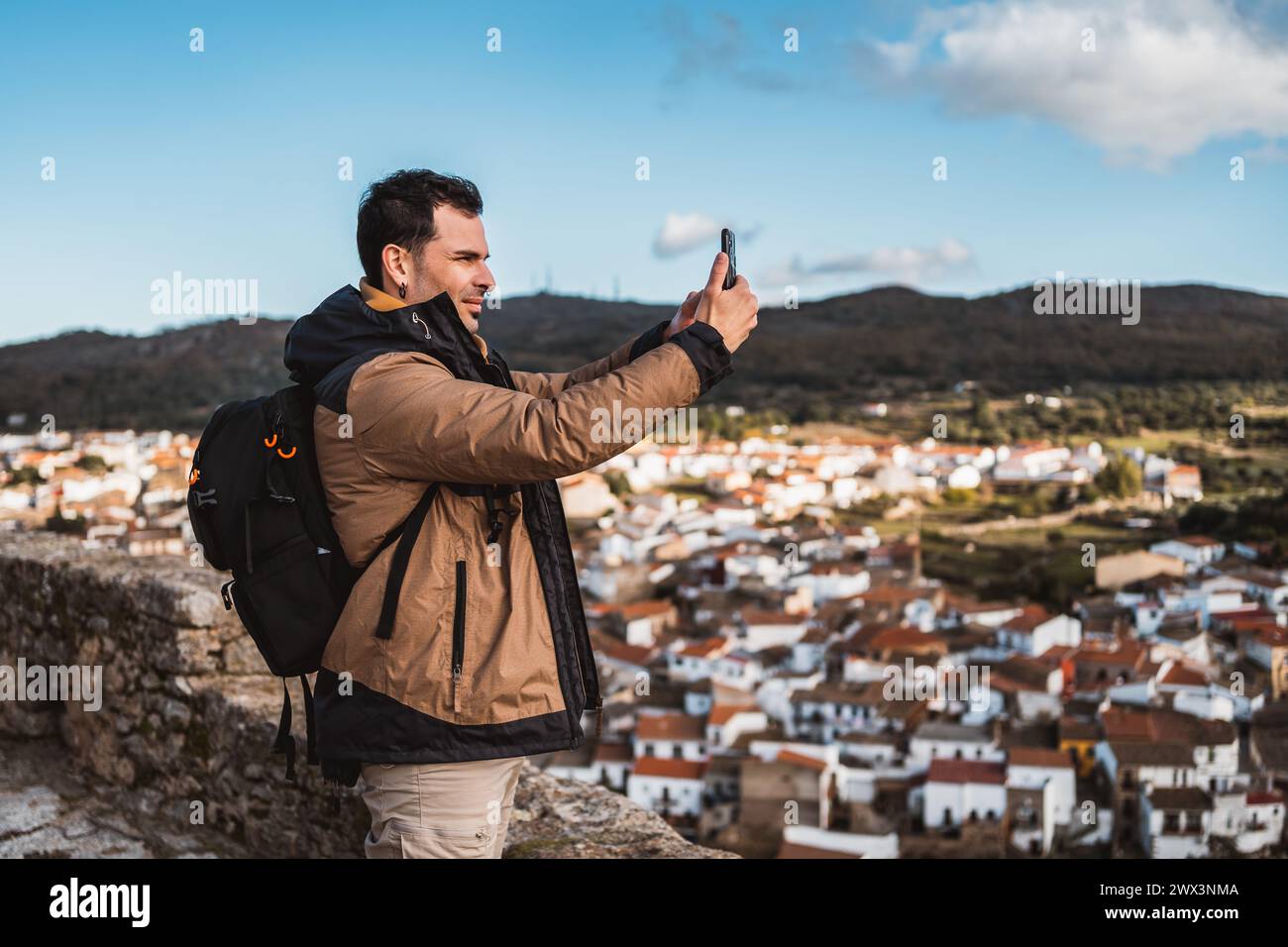 Young happy photographer man traveling sightseeing in a small town, taking a photo with his cell phone of the panoramic view of the town and the mount Stock Photo