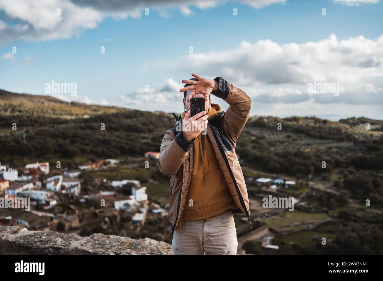 Young happy photographer man traveling sightseeing in a small town, taking a photo with his cell phone of the panoramic view of the town and the mount Stock Photo