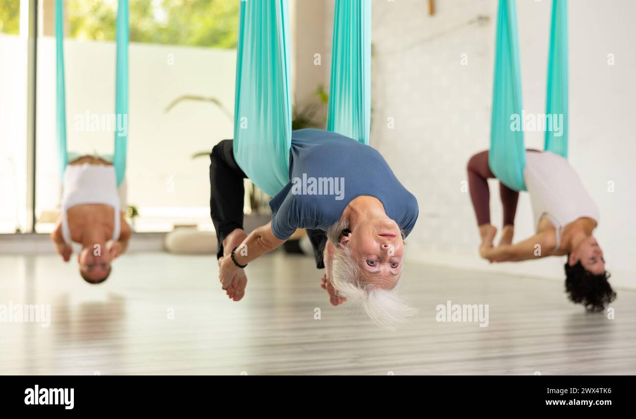 Senior woman practicing aerial yoga asana, hanging during group class in gym Stock Photo