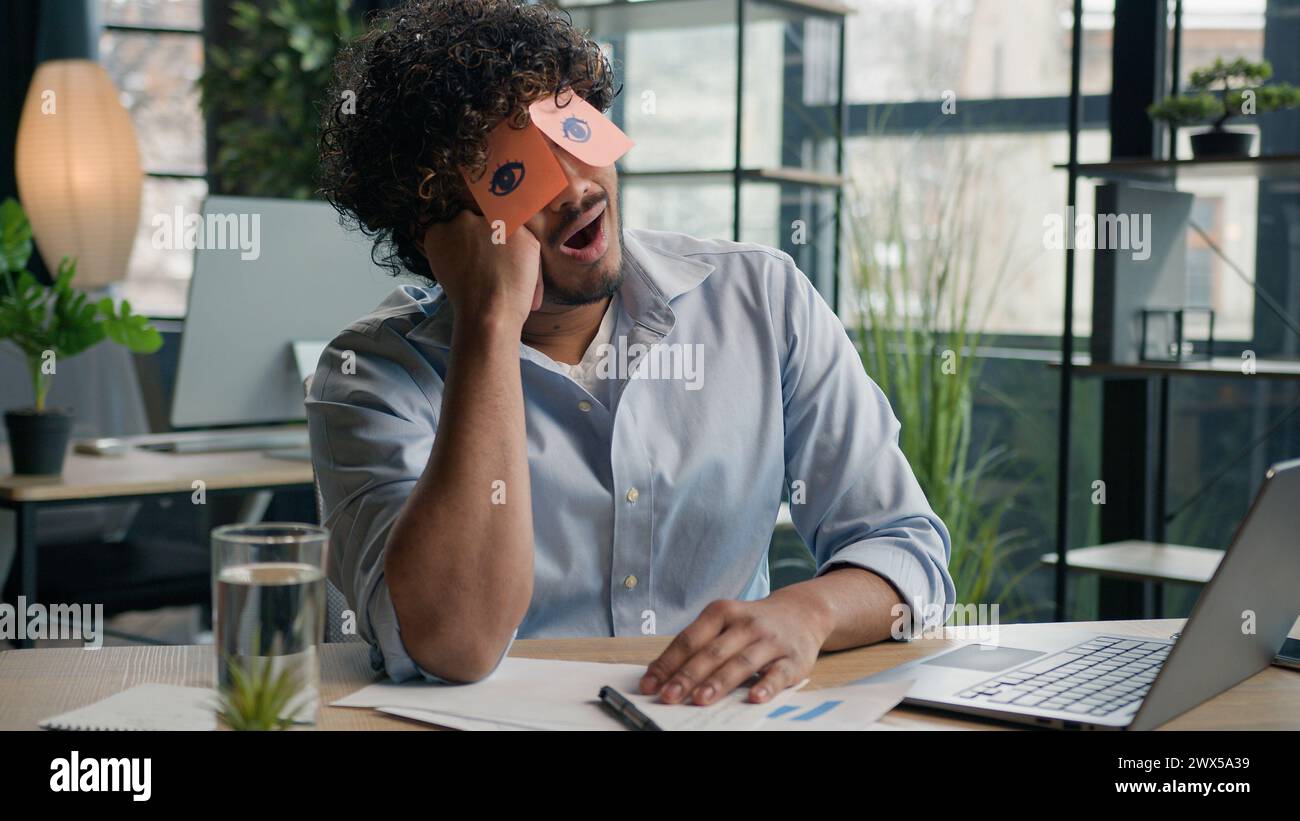 Funny lazy worker napping at office tired Arabian Indian business man sleeping covering eyes with sticky notes comical stickers on face employee Stock Photo
