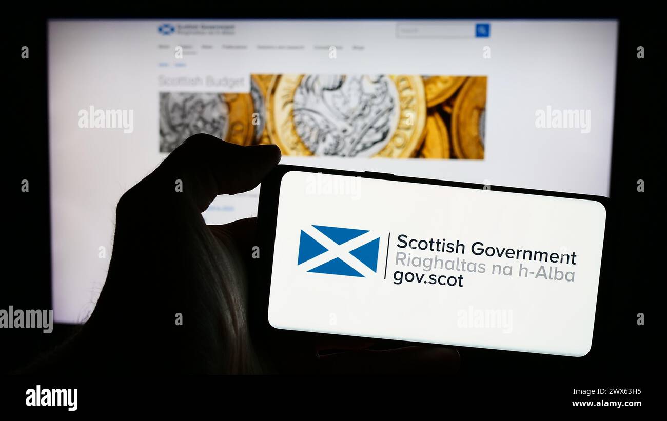 Person holding cellphone with logo of Scottish Government (Riaghaltas na h-Alba) in front of webpage. Focus on phone display. Stock Photo