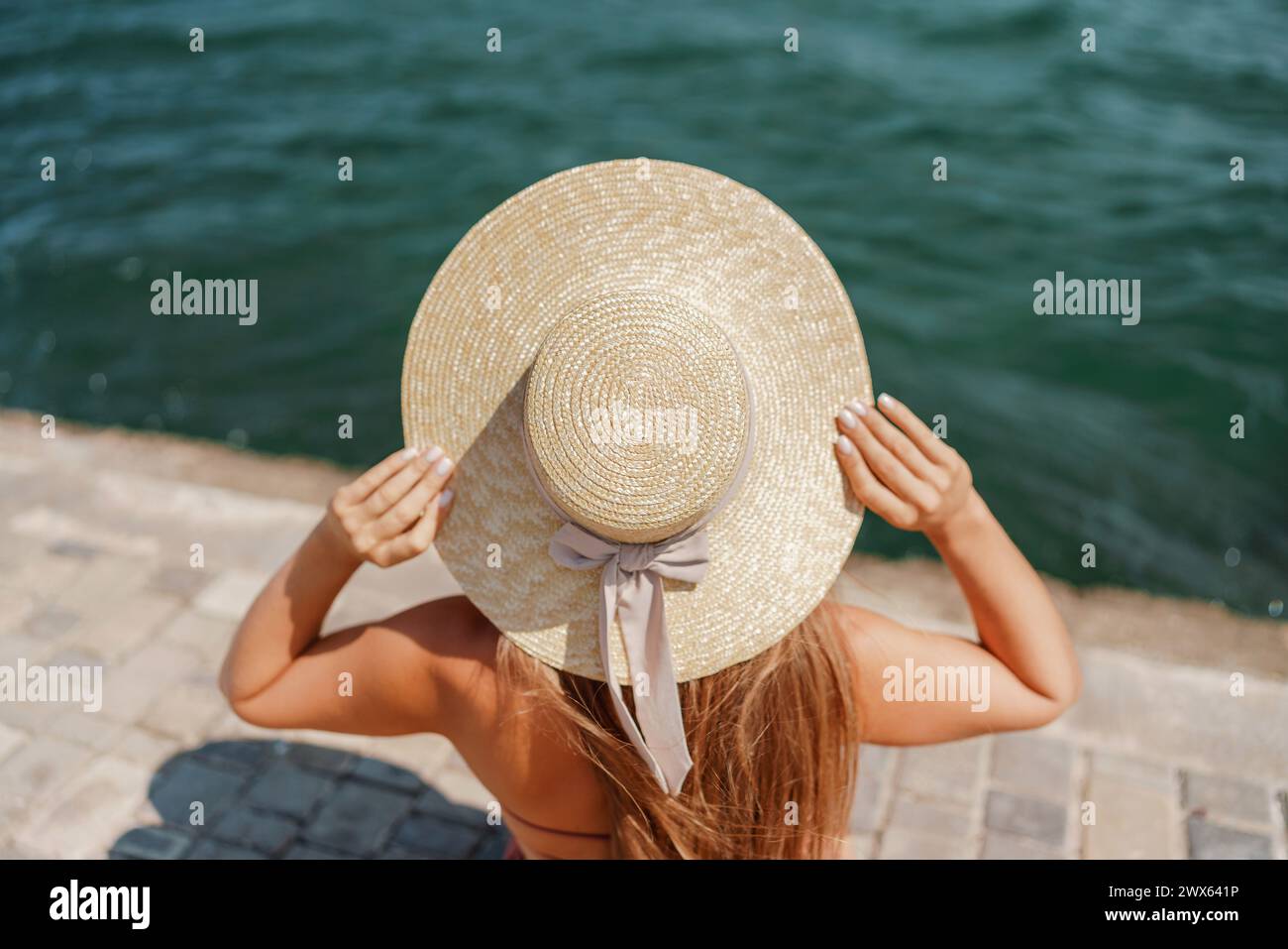 A woman in a swimsuit sits with her back holding a hat, looks at the ...