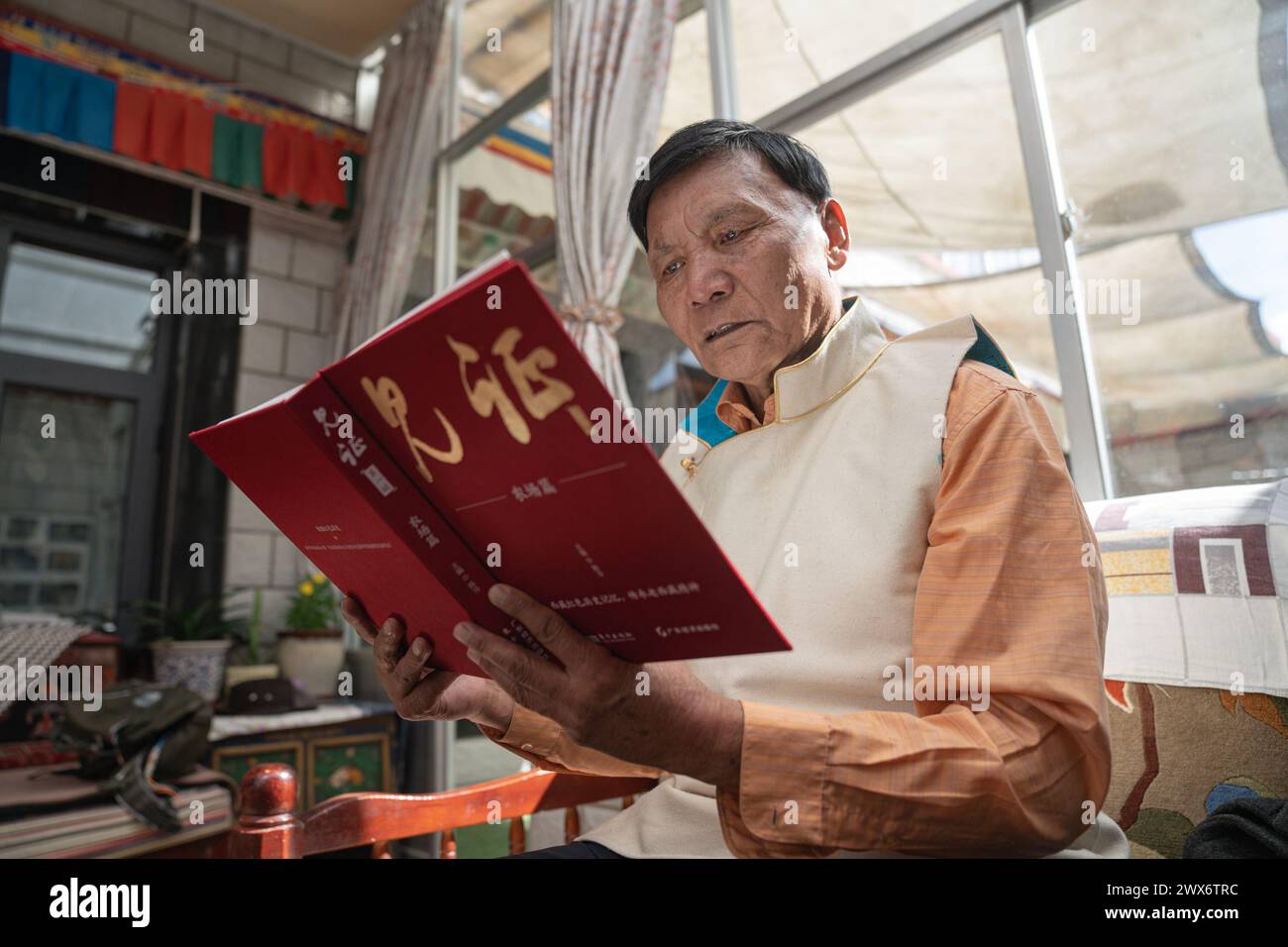 (240328) -- LHASA, March 28, 2024 (Xinhua) -- Phutsering reads a book at home in Lhasa, southwest China's Xizang Autonomous Region, March 23, 2024. Born in 1944 in Bainang County of Xigaze City, Phutsering was raised in a former serf family. A humble shack, situated near the manor where his mother toiled as a serf, served as his childhood home. Meanwhile, his father, employed at a separate manor, struggled to make ends meet by farming and crafting during lean months, barely generating sufficient income to cover the rent and taxes for their dwelling and fields. At the age of thirteen, he greete Stock Photo
