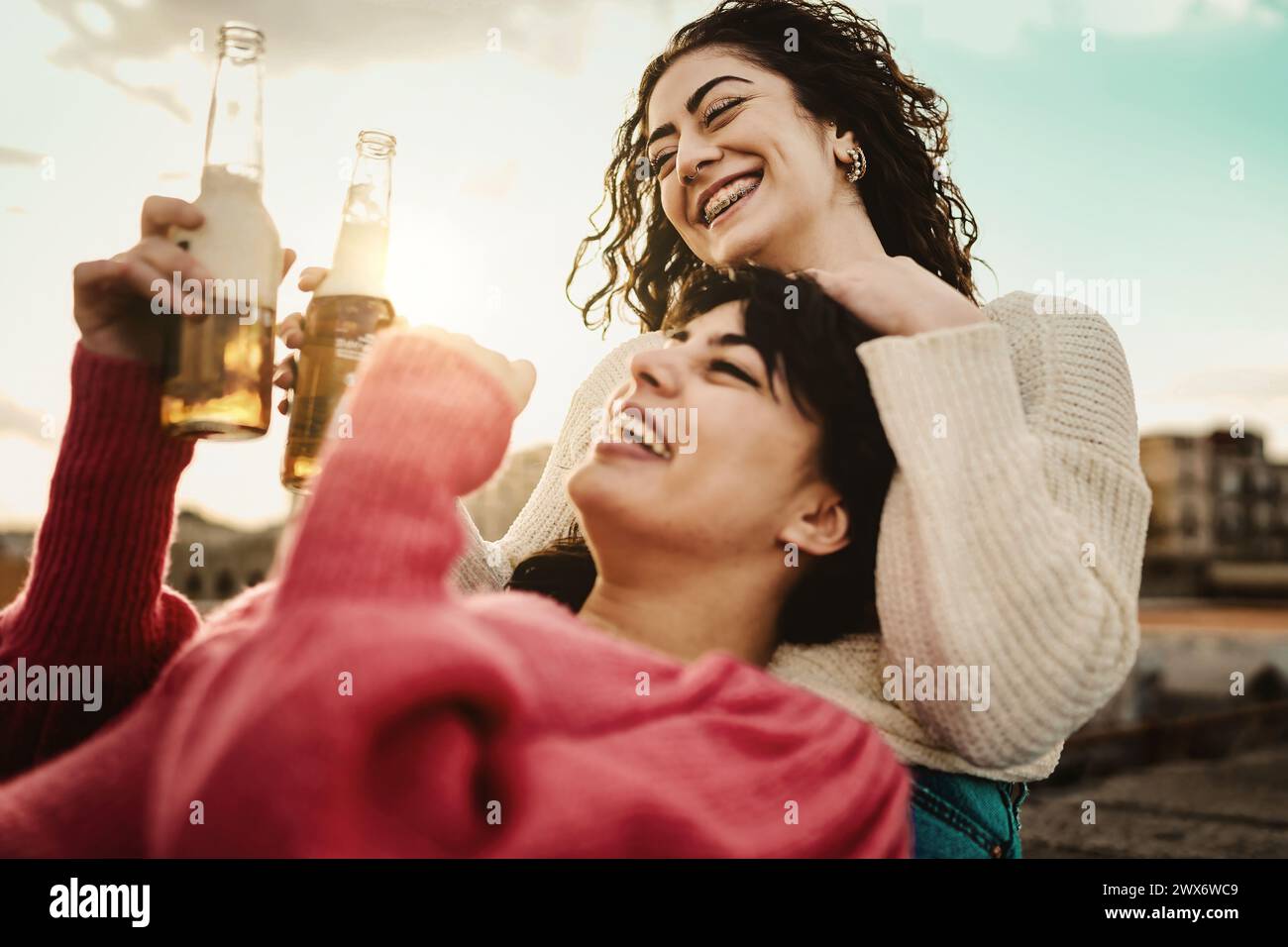 Two women laughing with beers - sunset celebration, golden hour warmth - carefree outdoor fun, candid happiness. Stock Photo